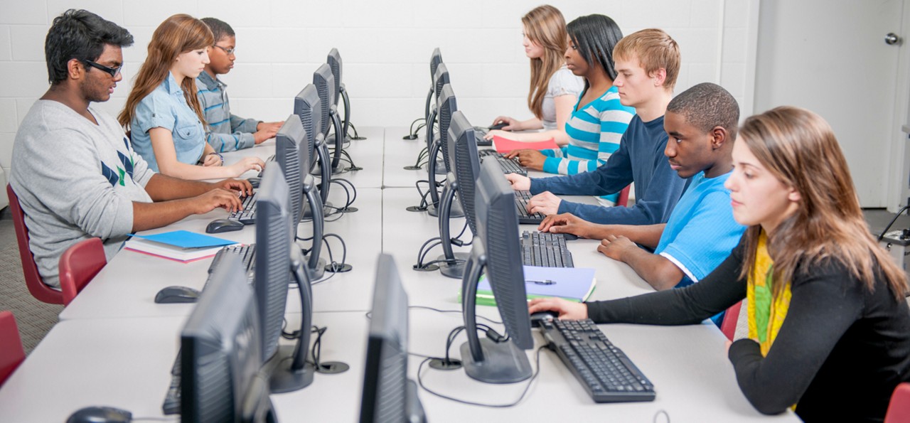 A multi-ethnic group of high school age students are sitting in a row at the computer lab and are doing research for an assignment online.